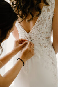 Tampa Modern Boho Bride Getting Wedding Ready with Bridesmaids Buttoning Open V Back with Delicate Floral Lace Wedding Dress