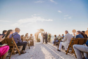 Beach Front Half Circle Arch Wedding Ceremony Couple Exchanging Vows Portrait | Wooden Folding Chairs and Pink Florals with Greenery