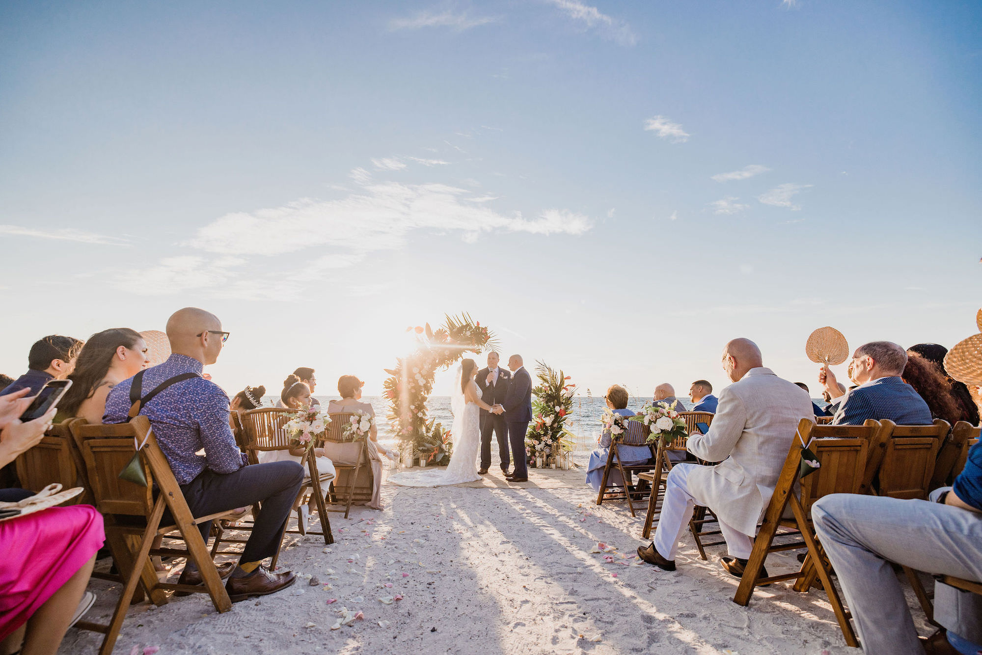 Beach Front Half Circle Arch Wedding Ceremony Couple Exchanging Vows Portrait | Wooden Folding Chairs and Pink Florals with Greenery
