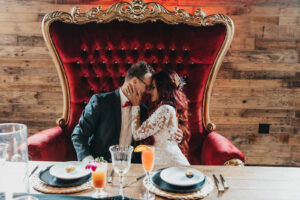 Wedding Couple Portrait Sitting in Modern Sweetheart Table with Red Velvet Loveseat Greenery and Floral Details | Gabro Event Rental Services