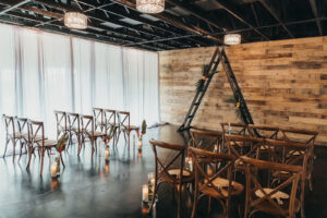Wooden Cross Back Chairs with Palm and Candle Aisle Décor Detail and Wooden Triangle Arch in Indoor Industrial Ceremony | Tampa Florida Wedding Rentals Gabro Event Services