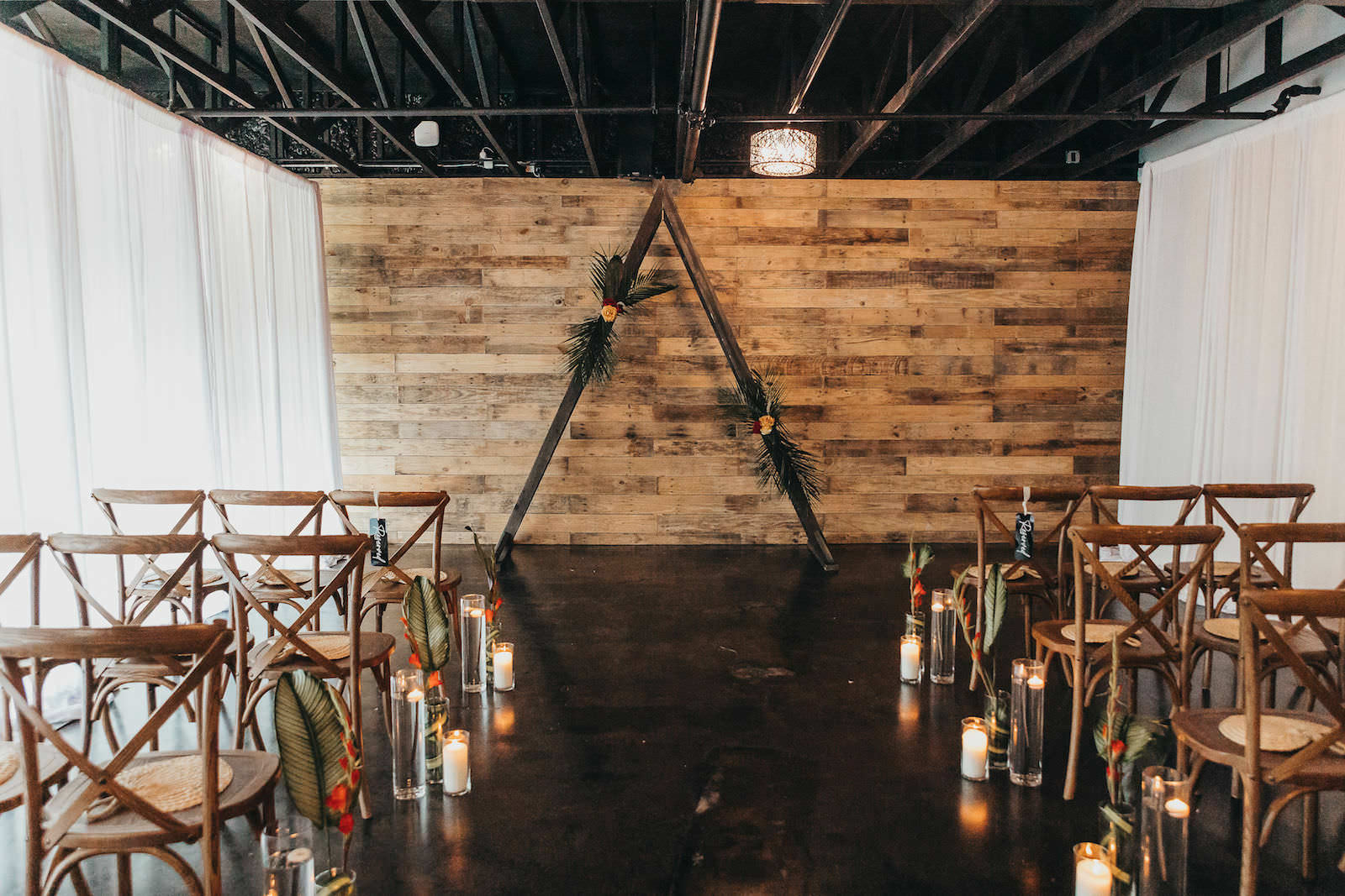 Wooden Cross Back Chairs with Palm and Candle Aisle Décor Detail and Wooden Triangle Arch in Indoor Industrial Ceremony | Tampa Florida Wedding Rentals Gabro Event Services