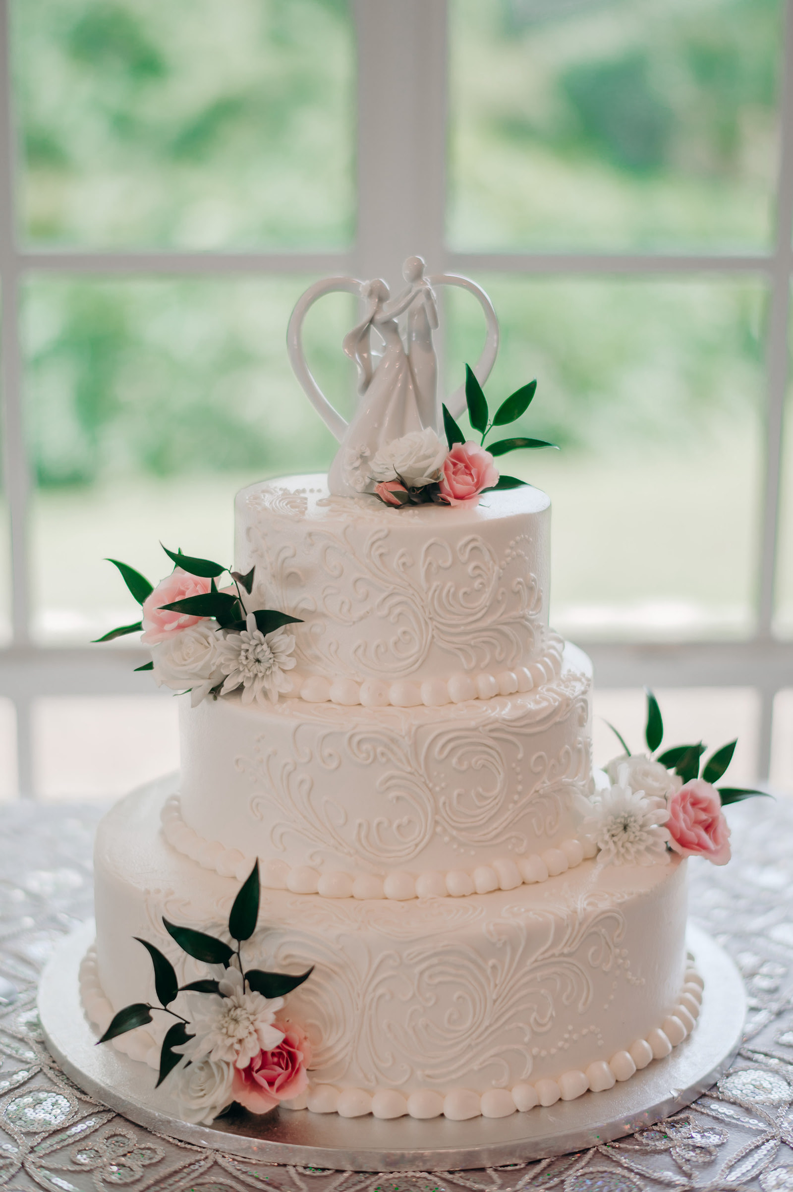 Three Tier White Detailed Cake with Pink, White, and Greenery Floral Detailing and Bride and Groom Cake Topper