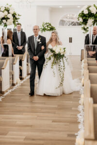 Winter Inspired Wedding Ceremony, Bride and Father Walking Down the Aisle Holding Lush White Flower and Greenery Hanging Floral Bouquet