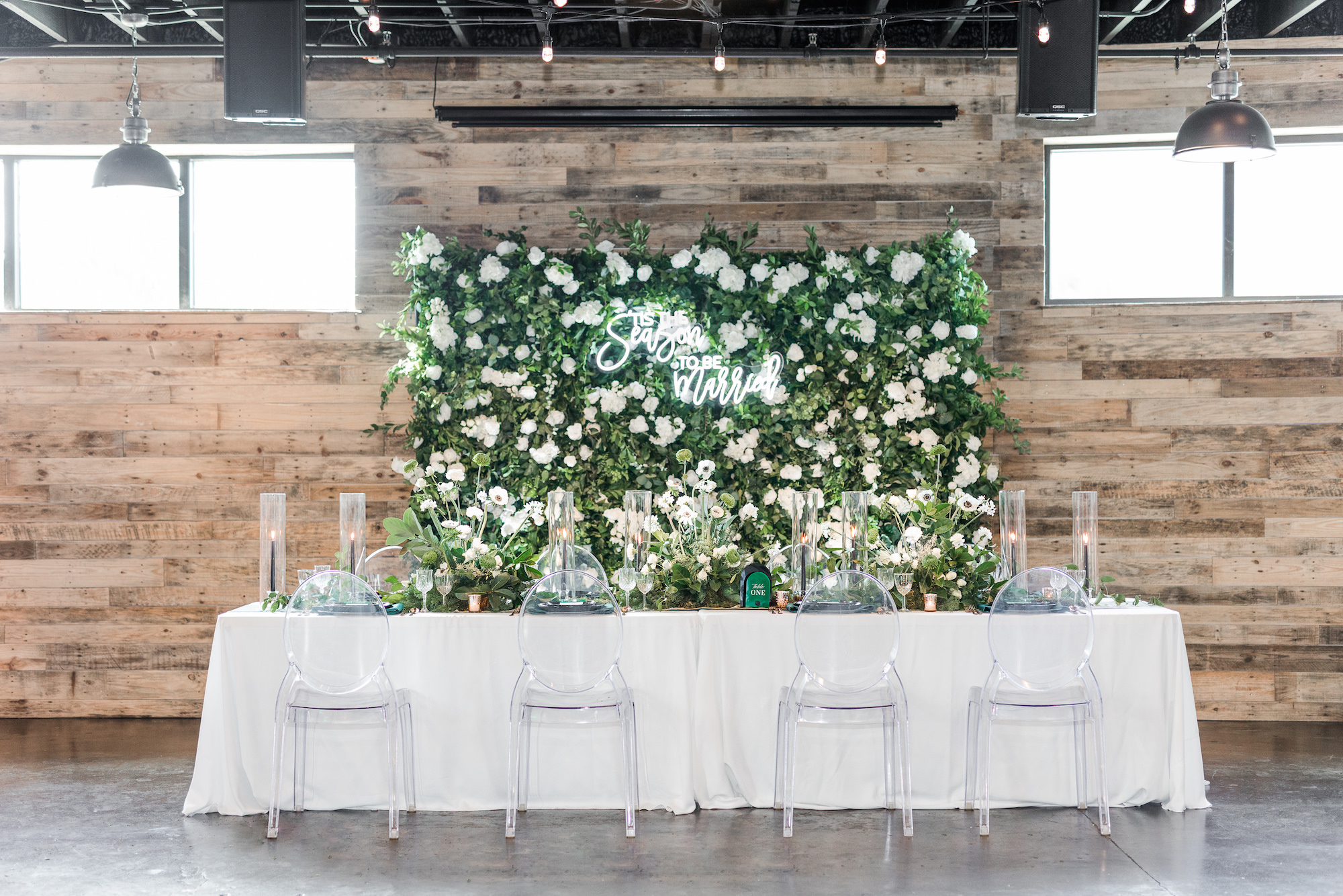 Winter Modern Whimsical Wedding Reception Decor, Long Feasting Table with White Linen, Acrylic Ghost Chairs, Tall Glass Cylinders with Black Candlesticks Greenery Wall with Lush White Roses, Hydrangeas and White "Tis the Season to Be Married" Neon Sign | Tampa Bay Wedding Planner MDP Events Planning | Madeira Beach Industrial Wedding Venue The West Events | Furniture and Linen Rentals Gabro Event Services