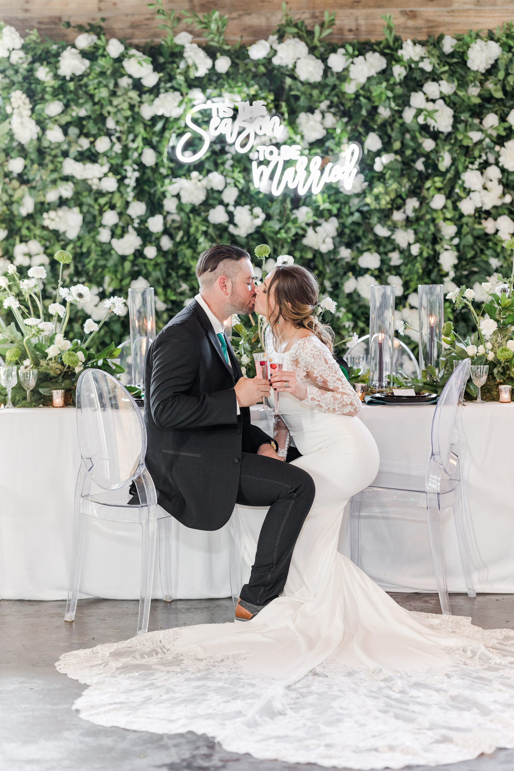 Winter Modern Whimsical Wedding Reception Styled Shoot Decor, Bride Wearing Sleek Lace and Illusion Long Sleeve Wedding Dress Kissing Groom Sitting in Ghost Acrylic Chairs, Long Feasting Table with White Table Linen, Lush Greenery and White Floral Wall Backdrop, White Neon Sign, Glass Cylinders with Black Candlesticks | Tampa Bay Wedding Planner MDP Events Planning | Wedding Furniture and Linens Gabro Event Services | Wedding Dress Truly Forever Bridal Tampa