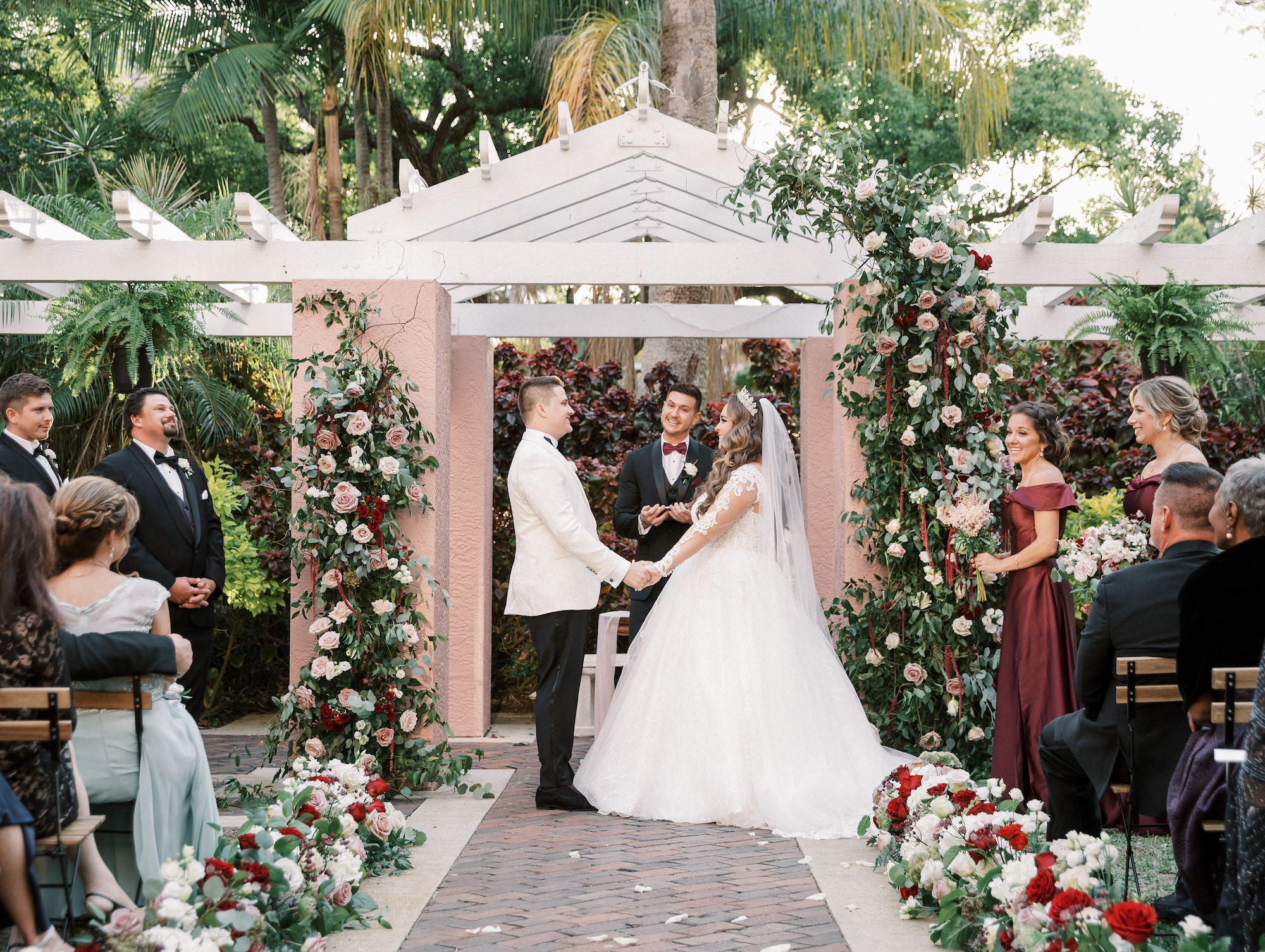 Elegant Royal Glam Gatsby Bride and Groom Exchanging Wedding Vows During Outdoor Courtyard Wedding Ceremony, Lush Greenery, Mauve, Blush Pink, Red and White Roses with Hanging Amaranthus Floral Pillars | Tampa Bay Wedding Planner and Designer John Campbell Weddings | St. Pete Wedding Venue The Vinoy Renaissance