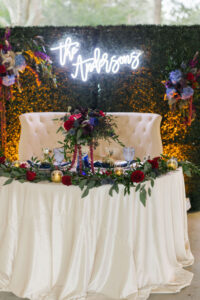 Couples Table with White Linen and White Sofa and Greenery Décor with Indigo and Hot Pink Florals and a Neon Sign on the Greenery Wall | Tampa Bay Rentals Kate Ryan Event Rentals