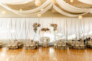 Fall Boho Wedding Ballroom Reception Decor, Long Feasting Table with Taupe Table Linens, Gold Chiavari Chairs, Lush Fall Colored Floral Arrangements, Drape Ceiling White Linens, String Lights, Rattan Chandeliers | Tampa Bay Wedding Rentals Gabro Event Services