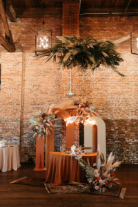 Earthy Neutral Boho Modern Chic Wedding Reception Decor, Sweetheart Table with Terracotta Table Linen, Cream and Brown Arched Panels, Lush Floral Bouquets with Pampas Grass, Monstera Palm Leaves, Palm Leaves Greenery with Pampas Grass Floral Chandelier | Tampa Bay Wedding Planner Wilder Mind Events | Wedding Florist Save the Date Florida | Wedding Venue Armature Works
