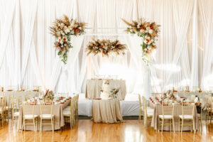 Fall Boho Wedding Ballroom Reception Decor, Long Feasting Table with Taupe Table Linens, Gold Chiavari Chairs, Lush Fall Colored Floral Arrangements, Drape Ceiling White Linens, String Lights, Rattan Chandeliers | Tampa Bay Wedding Rentals Gabro Event Services