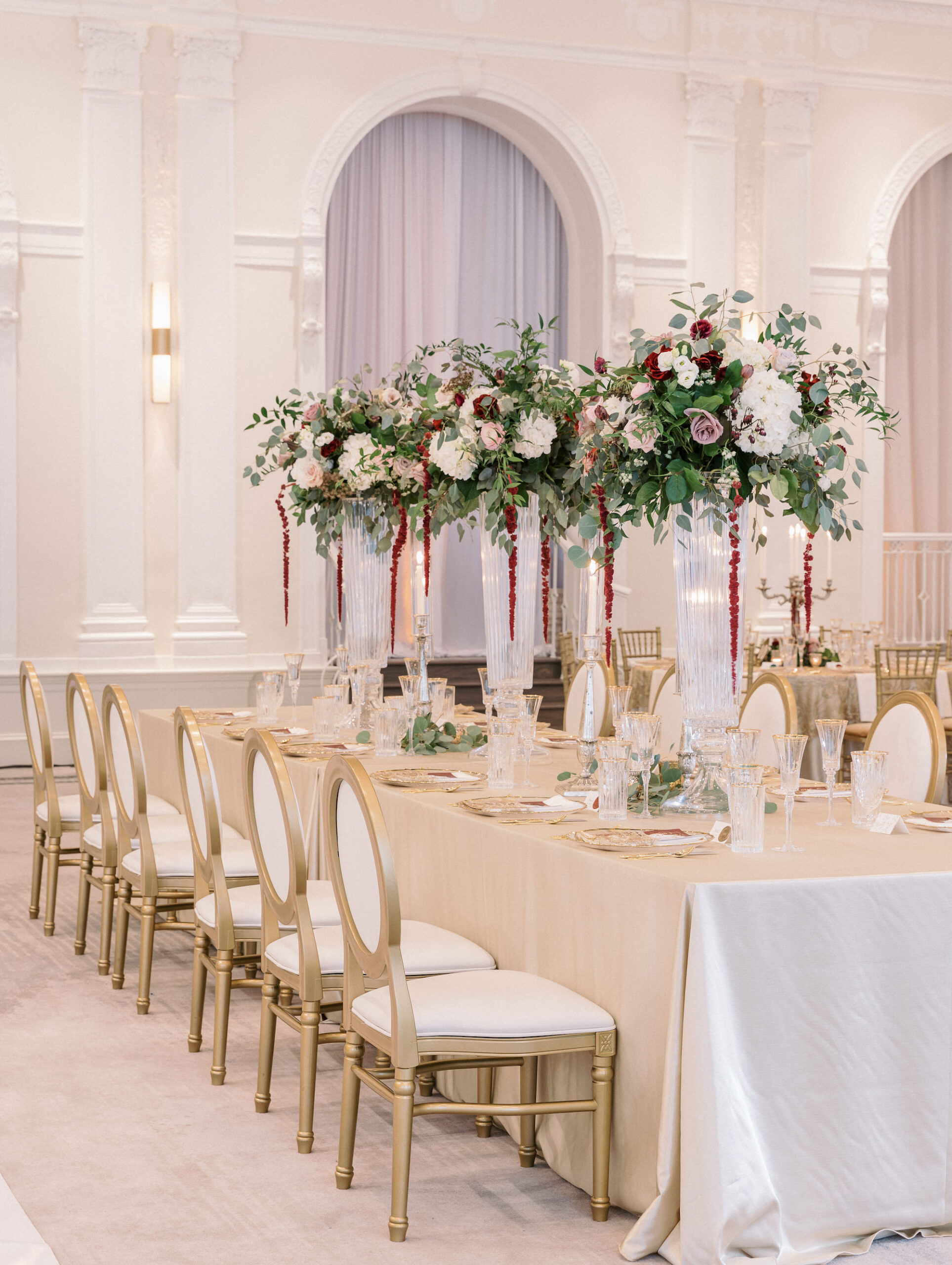 Elegant Royal Glam Gatsby Wedding Reception Decor, Long Table with Champagne Linen, Gold and Ivory Dining Chairs, Tall Glass Vases with Lush Greenery, White, Mauve, Burgundy and Blush Pink Roses with Red Hanging Amaranthus Flower Centerpieces | Tampa Bay Wedding Planner and Designer John Campbell Weddings | St. Pete Wedding Rentals Kate Ryan Event Rentals
