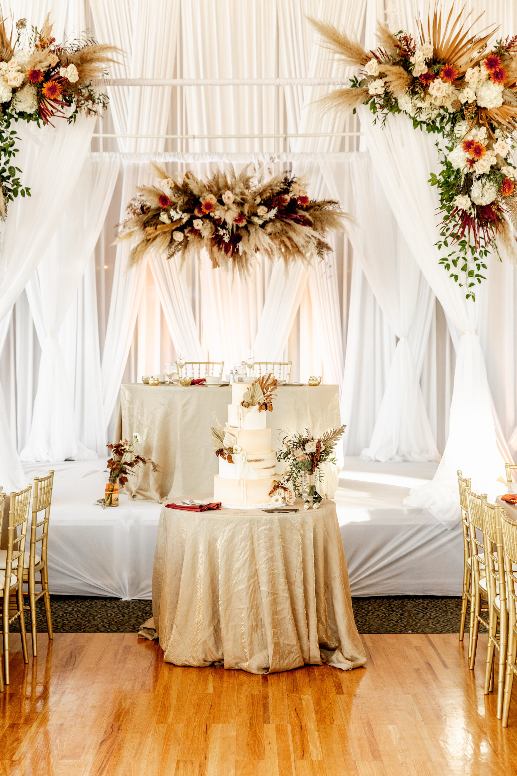 Fall Boho Wedding Ceremony Decor, Cake Table with Champagne Linen and Five Tier White Wedding Cake | Tampa Bay Wedding Rentals Gabro Event ServicesArch with Lush Dried Palm Leaves, Pampas Grass, Burnt Orange, White and Ivory Flowers, Greenery Floral Arrangements