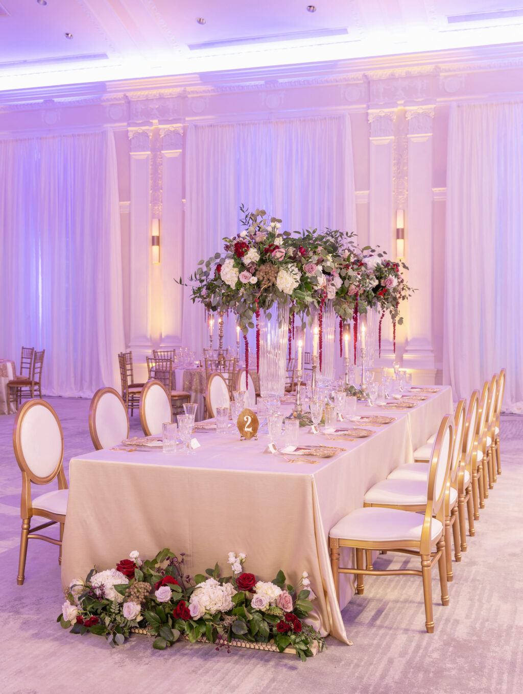 Elegant Royal Glam Gatsby Wedding Reception Decor, Purple Uplighting, Long Table with Champagne Linen, Gold and Ivory Dining Chairs, Tall Glass Vases with Lush Greenery, White, Mauve, Burgundy and Blush Pink Roses with Red Hanging Amaranthus Flower Centerpieces | Tampa Bay Wedding Planner and Designer John Campbell Weddings | St. Pete Wedding Rentals Kate Ryan Event Rentals