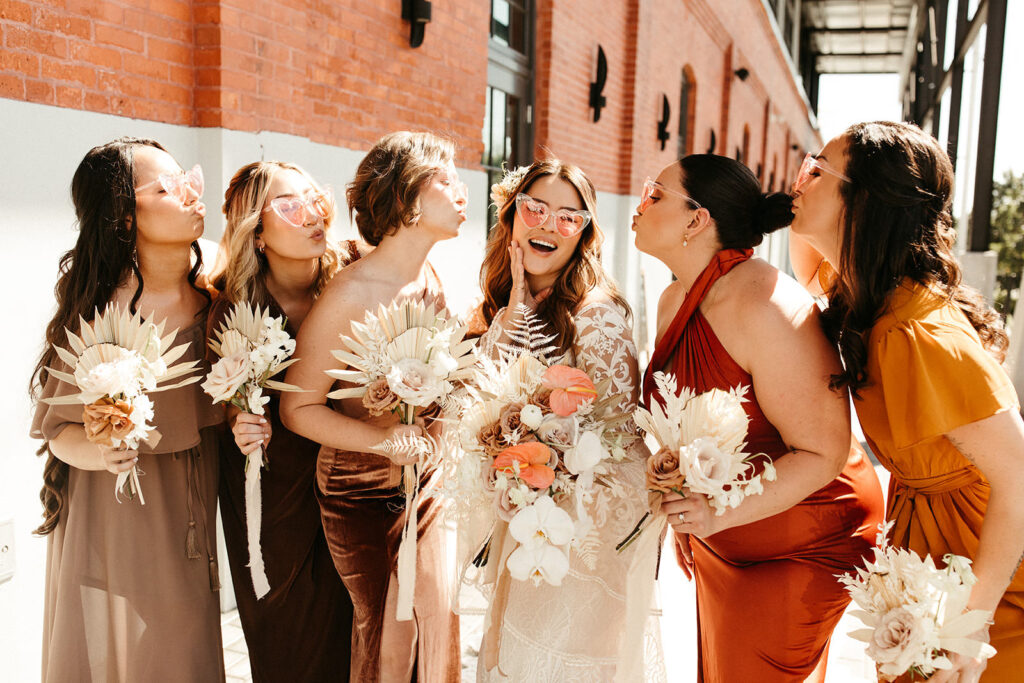 Earthy Neutral Boho Modern Chic Bridal Party, Bride Wearing Clear and Pink Heart Lens Sunglasses Holding White Orchids, Pink Anthuriums, Dried Leaves Floral Bouquet, Bridesmaids Wearing Mix and Match Terracotta, Gold and Bronze Dresses | Tampa Bay Wedding Venue Armature Works | Wedding Florist Save the Date Florida | Wilder Mind Events