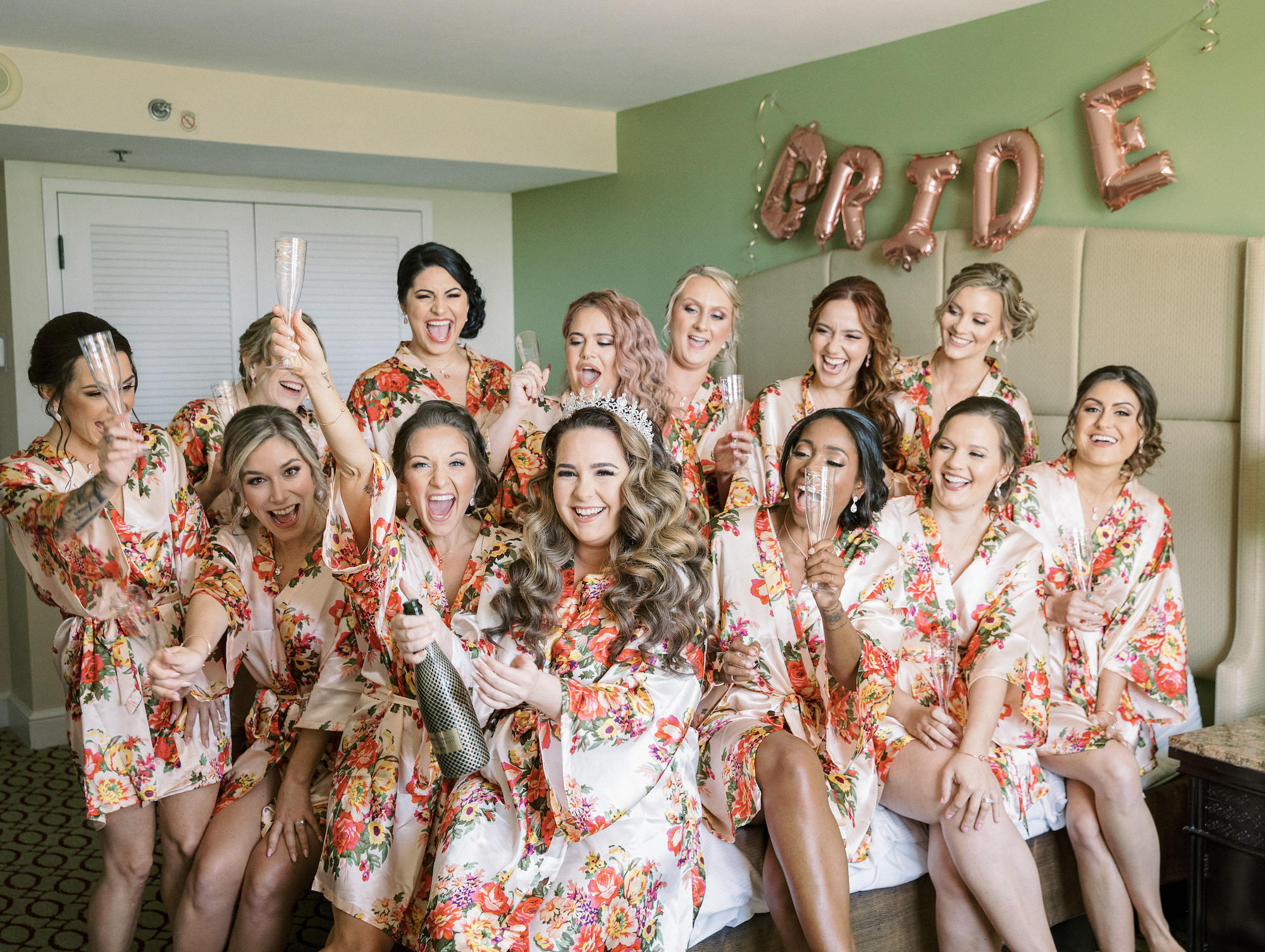 Royal Glam Gatsby Wedding, Bridal Party Getting Wedding Ready, Bridesmaids in Pink Floral Robes Popping Champagne with Bride in Princess Tiara | Tampa Bay Wedding Hair and Makeup Femme Akoi Beauty Studio