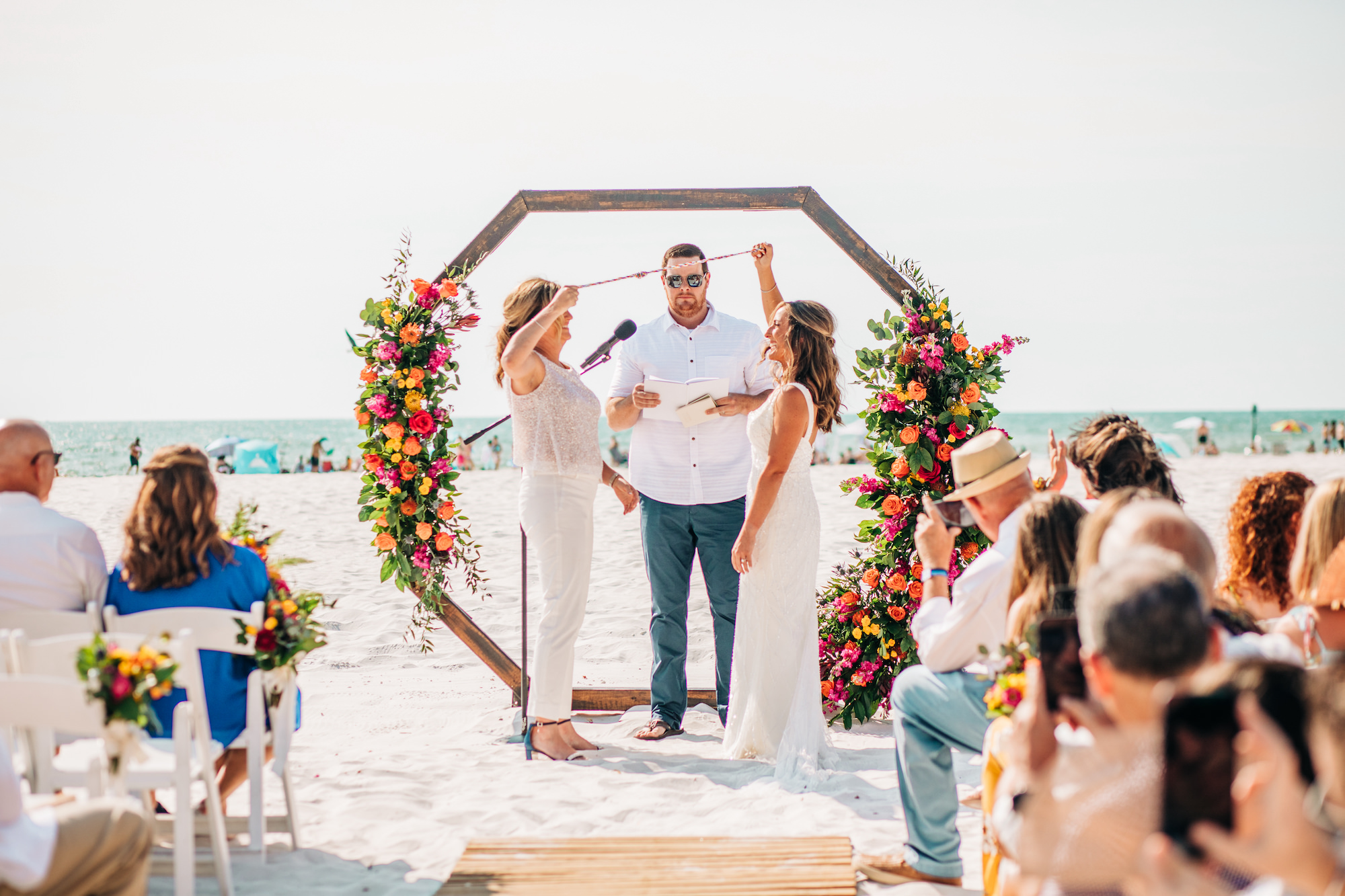 Vibrant Colorful Same Sex Wedding Ceremony, Brides Doing Unity Ceremony, Geometric Wooden Arch with Lush Yellow, Pink, Orange and Greenery Floral Arrangements | Waterfront Wedding Venue Hilton Clearwater Beach