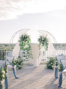 Two Half Circle Arches with Greenery and White Floral Details | White Ceremony Chairs with Blue Draping and White Floral and Greenery Detailing | Tampa Wedding Rentals Gabro Event Services