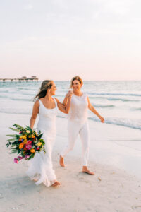Vibrant Colorful Same Sex Wedding, Brides Walking on Clearwater Beach Holding Lush Greenery, Pink, Yellow and Orange Flower Bouquet