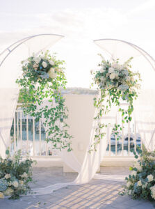 Two Half Circle Arches with Greenery and White Floral Details | White Ceremony Chairs with Blue Draping and White Floral and Greenery Detailing | Tampa Wedding Rentals Gabro Event Services