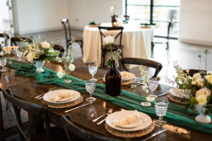 Modern Whimsical Wedding Reception Decor, Long Wooden Feasting Table with Emerald Green Cheesecloth Table Runner, Rattan Round Place Mats, Simple White and Greenery Floral Centerpieces, Wooden Cross Back Chairs | Unique Tampa Wedding Venue Florida Ave Brewing