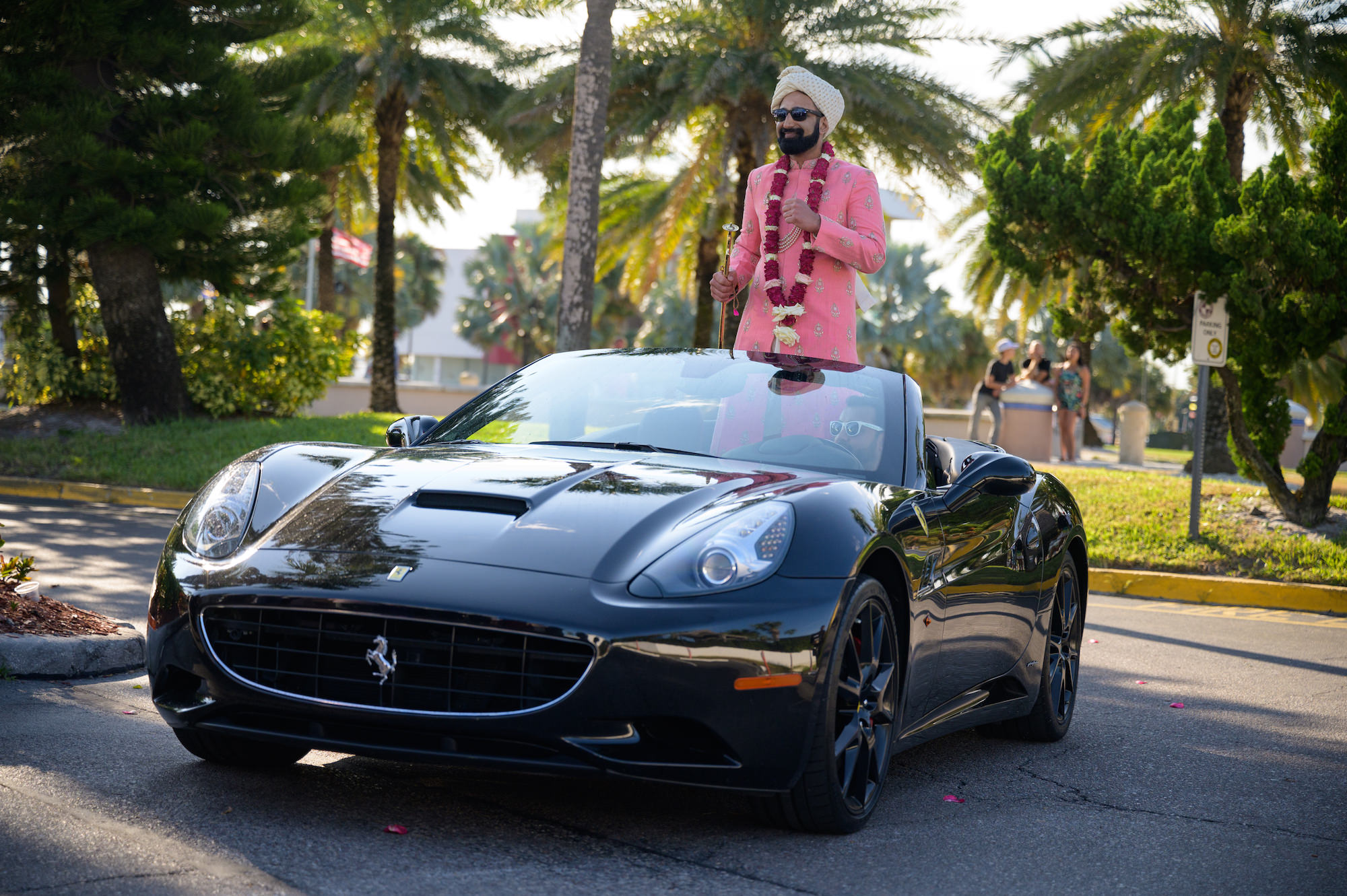 Florida Groom Baraat Arrival in Black Ferrari Convertible Luxury Sports Car, Groom Wearing Traditional Indian Wedding Kurta in Bright Pink and Gold, Red Hindu Garland