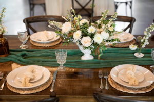 Modern Whimsical Wedding Reception Decor, Long Wooden Feasting Table with Emerald Green Cheesecloth Table Runner, Rattan Round Place Mats, Simple White and Greenery Floral Centerpieces, Wooden Cross Back Chairs | Unique Tampa Wedding Venue Florida Ave Brewing
