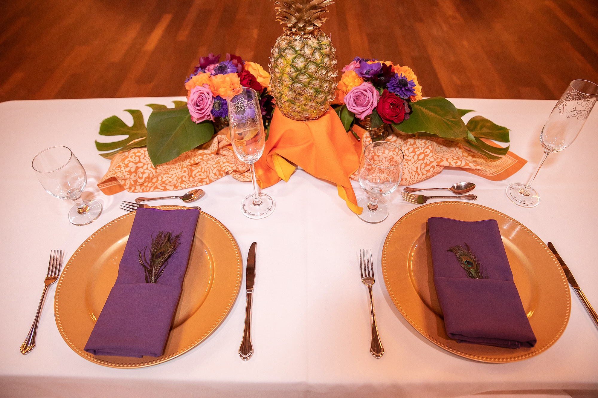 White Linen Tablescape with Gold Chargers Purple Napkins and Peacock Feather Detail and Pineapple and Tropical Floral Centerpieces