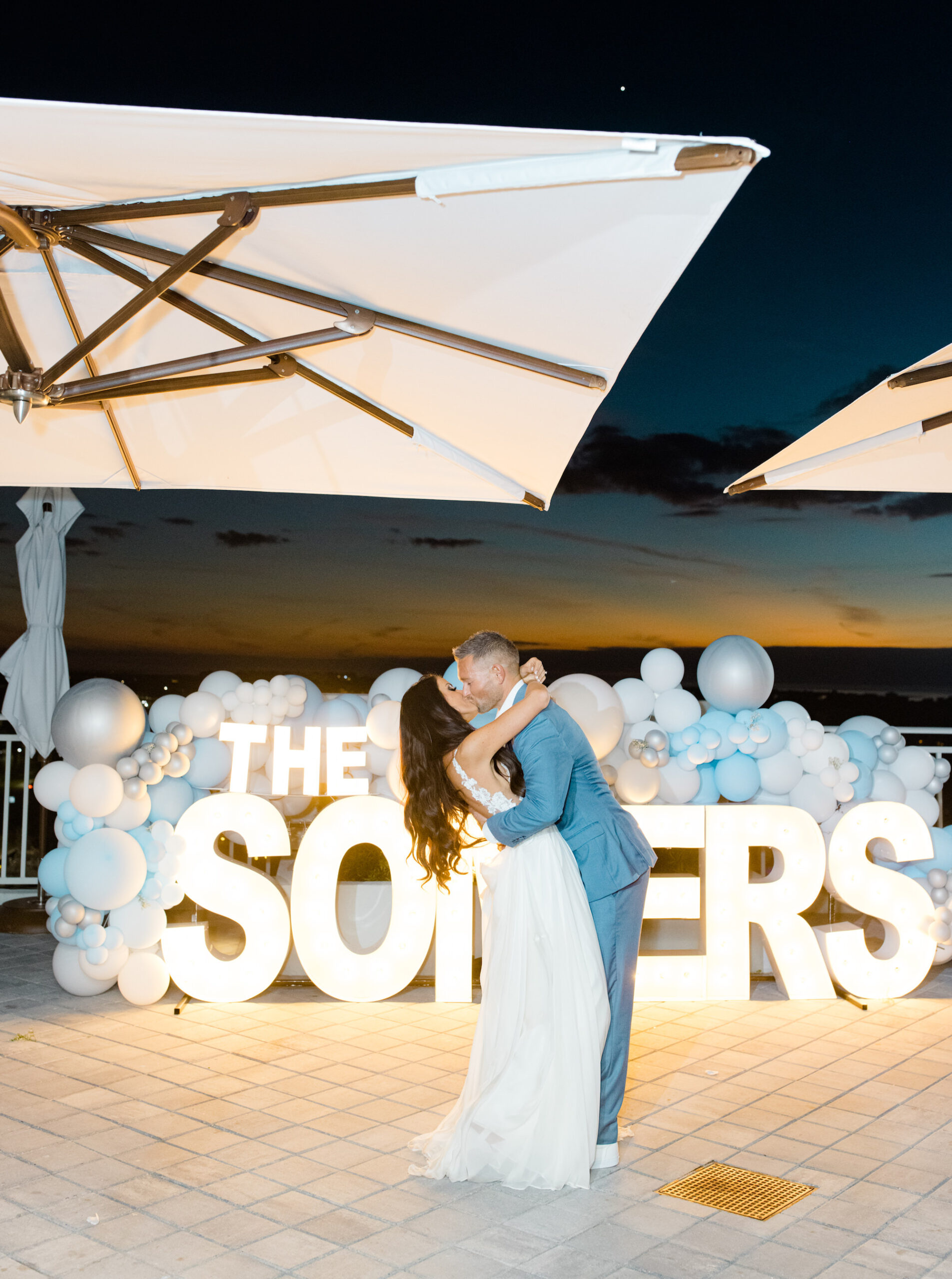 Bride and Groom Wedding Portrait Kissing In Front of White Light Up Marquee Letters with Blue, Silver, and White Balloons