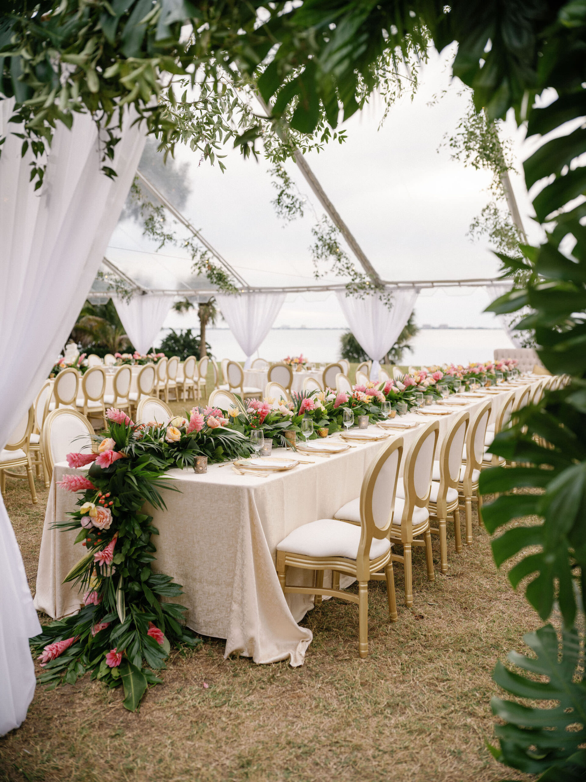 Luxurious Classic Wedding Tent Reception Decor with Lush Monstera Palm Leaves Arch, Long Feasting Table with Ivory Linens and Gold and Cream Dining Chairs, Monstera Palm Leaves with Pink Ginger Tropical Flowers and Peach Roses Garland | Tampa Bay Wedding Florist Botanica International Design Studio