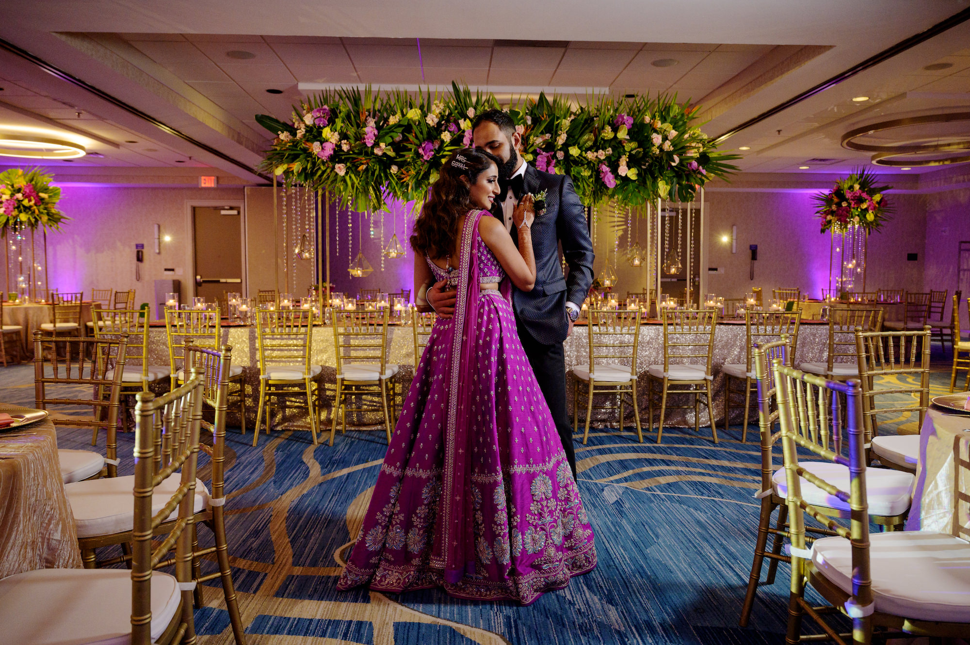 Indian Bride and Groom at Reception, Luxurious Decor with Head Feasting Table Adorned with Tall Floral Centerpieces, Tropical Flowers, Gold Chiavari Chairs, Bride Wearing Traditional Indian Fuchsia Lehenga and Groom Wearing Modern Black Tuxedo | Tampa Bay Hair and Makeup Artist Michelle Renee the Studio | Gabro Event Rentals | Hilton Clearwater Beach Resort and Spa