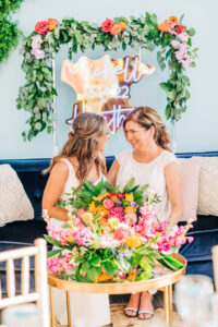 Vibrant Colorful Same Sex Wedding, Brides Sitting on Blue Velvet Couch with Lush Greenery, Pink, Yellow, Orange Floral Bouquet, Neon Custom Sign Backdrop | Tampa Bay Wedding Rentals Kate Ryan Event Rentals, A Chair Affair