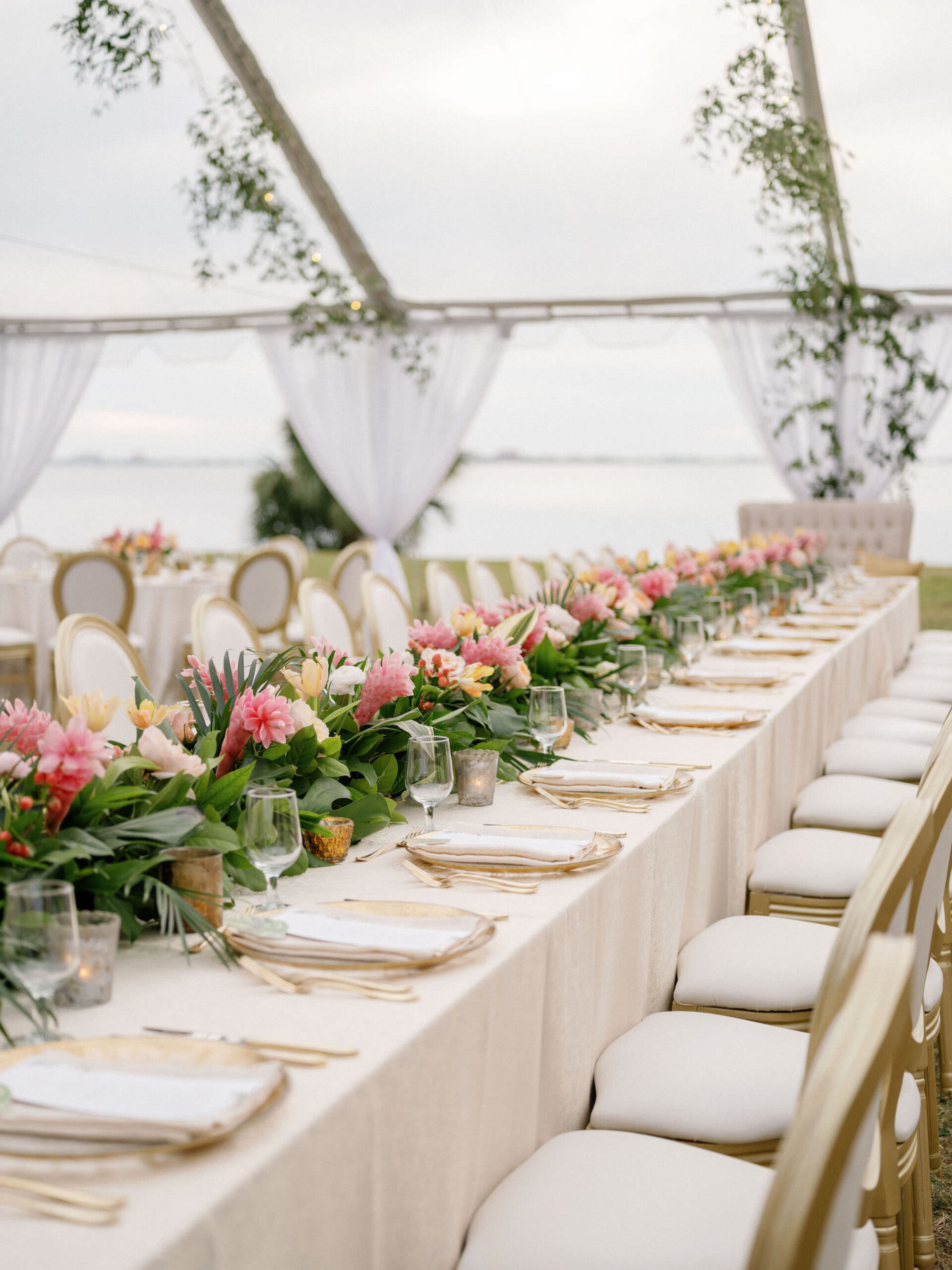 Luxurious Classic Wedding Reception Decor, Long Feasting Table with Ivory Linens, Gold and Cream Dining Chairs, Gold Flatware, Lush Monstera Palm Leaves with Pink Ginger Tropical Flowers | Tampa Bay Wedding Florist Botanica International Design Studio