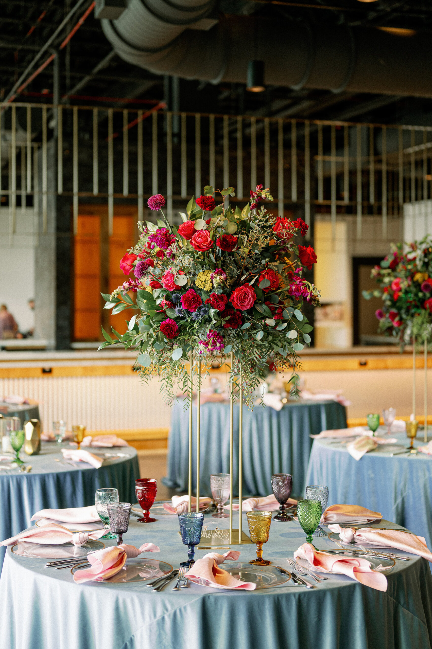 Vibrant Wedding Reception Decor, Round Tables with Dusty Blue Table Linens, Colorful Water Goblets, Tall Gold Stand with Lush Pink, Red Roses, Yellow Fuschia Flowers and Eucalyptus Greenery Leaves Centerpiece | Tampa Bay Wedding Photographer Dewitt for Love Photography | Wedding Rentals Gabro Event Services