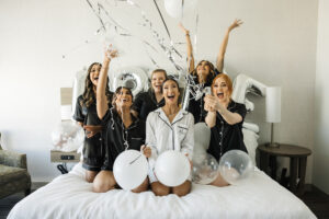 Bride and Bridesmaids in Matching Black Short Pajama Set on Bed with Silver Confetti Poppers and Balloons | Tampa Bay Wedding Hair and Makeup Femme Akoi Beauty Studio