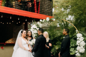 Bride and Groom Exchanging Wedding Vows, Simple Classic Outdoor Bamboo Courtyard Wedding Decor, Groom Wearing All Black Tuxedo, Bridesmaid in Elegant Black Dress, Hanging Bistro Lights, Round Circular Gold Metal Arch with Lush White and Greenery Flowers | Tampa Bay Wedding Photographer Amber McWhorter Photography | St. Pete Wedding Florist Save the Date Florist | Wedding Venue NOVA 535 | Wedding Officiant Brandi Morris