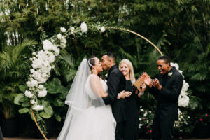 Bride and Groom Exchanging Wedding Vows, Simple Classic Outdoor Bamboo Courtyard Wedding Decor, Groom Wearing All Black Tuxedo, Bridesmaid in Elegant Black Dress, Hanging Bistro Lights, Round Circular Gold Metal Arch with Lush White and Greenery Flowers | Tampa Bay Wedding Photographer Amber McWhorter Photography | St. Pete Wedding Florist Save the Date Florist | Wedding Venue NOVA 535 | Wedding Officiant Brandi Morris