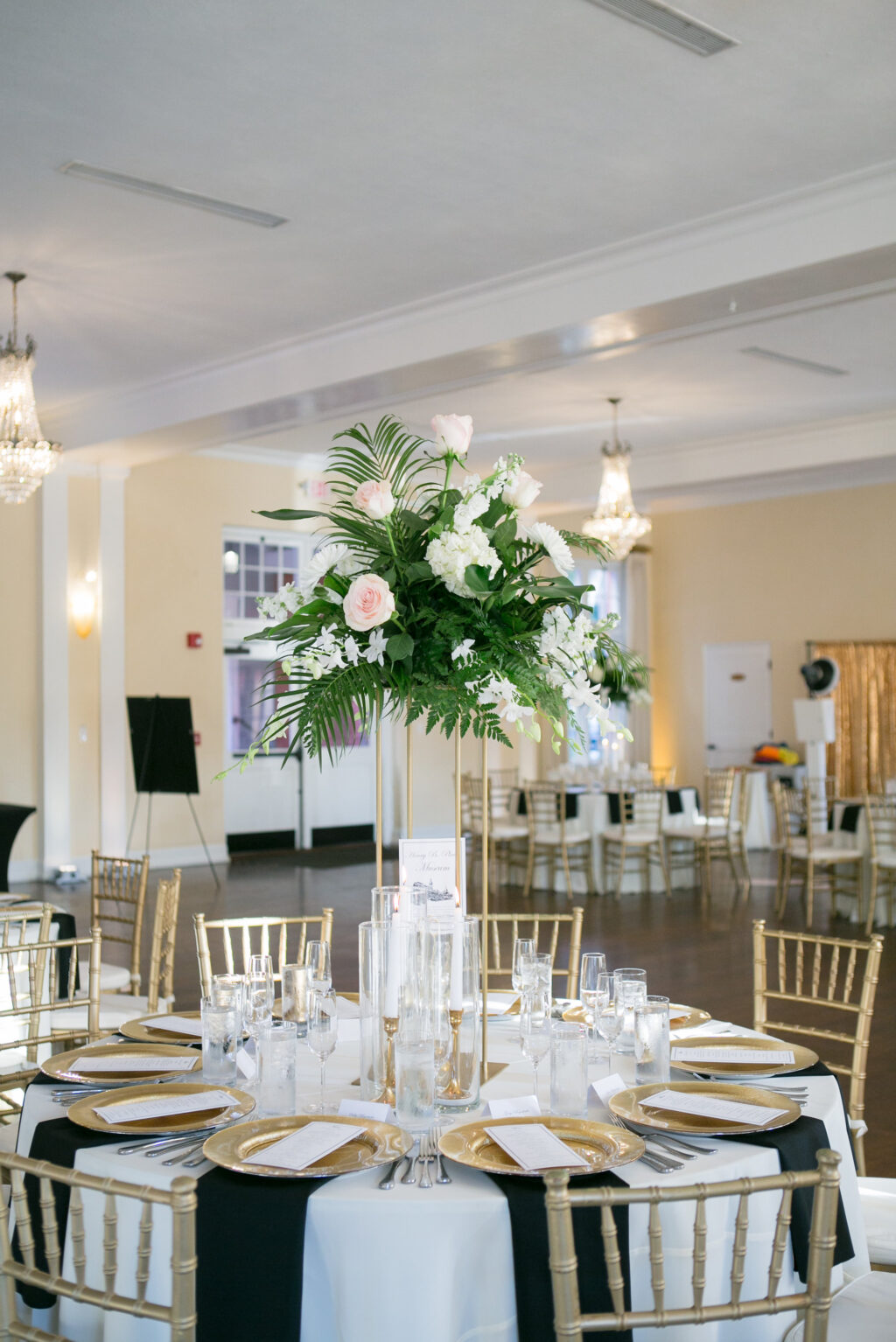 Elegant Tall Gold Flower Stand with White Tropical Flowers and Greenery Inspiration | Gold Chiavari Chairs | Ivory and Black Linens with Gold Chargers and Silver Flatware Wedding Reception Table Setting Ideas | Tampa Bay Rentals A Chair Affair