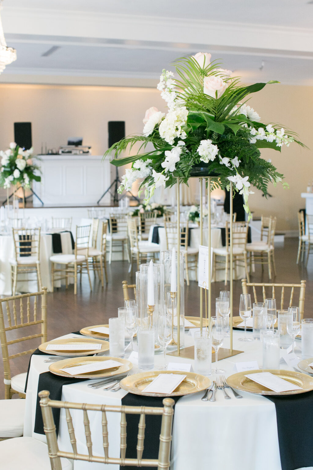 Elegant Tropical Tall Gold Flower Stand with White Tropical Flowers and Greenery Inspiration | Gold Chiavari Chairs | Ivory and Black Linens with Gold Chargers and Silver Flatware Wedding Reception Table Setting Ideas | Tampa Bay Rentals A Chair Affair