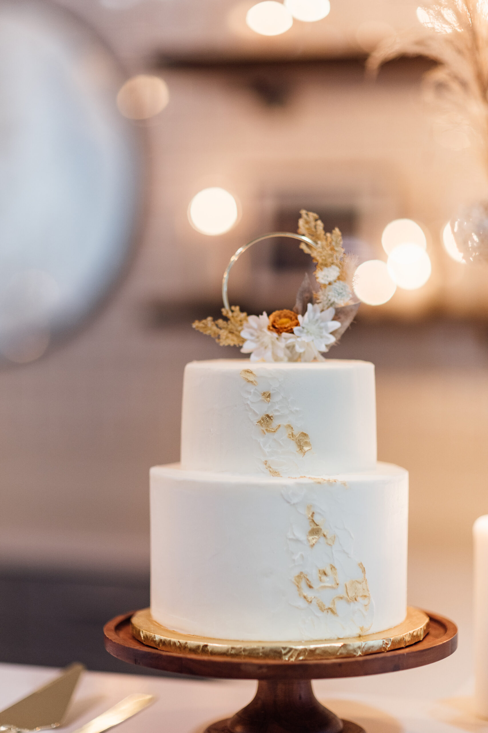 Modern Two-Tiered Smooth Buttercream White Wedding Cake with Gold Flakes and Boho Round Wedding Topper