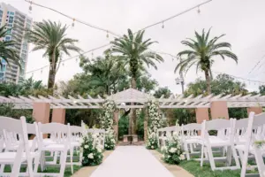 Classic Outdoor Courtyard Wedding with White Floral and Greenery Arch and Aisle Decor and White Folding Chair Wedding Ceremony Inspiration | St Pete Venue The Vinoy