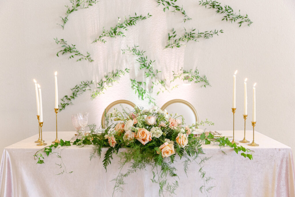 Sweetheart Table with Blush Pink Tablecloth Linen, Taper Candles and Gold Candle Holders | Peach and Pink Roses with Greenery Tabletop Flower Arrangement and Backdrop Wedding Reception Decor Inspiration