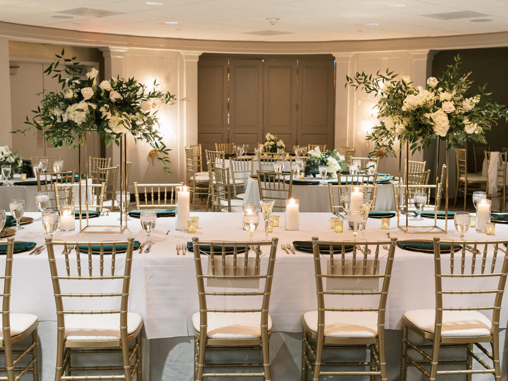 White Rose and Ruscus Greenery in Tall Gold Metal Geometric Column Flower Stand Centerpiece Inspiration | Canopy Ballroom Elegant Candlelit Wedding Reception Decor Ideas | Gold Mercury Votives | Gold Chiavari Chairs | Downtown Venue The Tampa Club | Over The Top Linen Rentals | Planner Perfecting The Plan