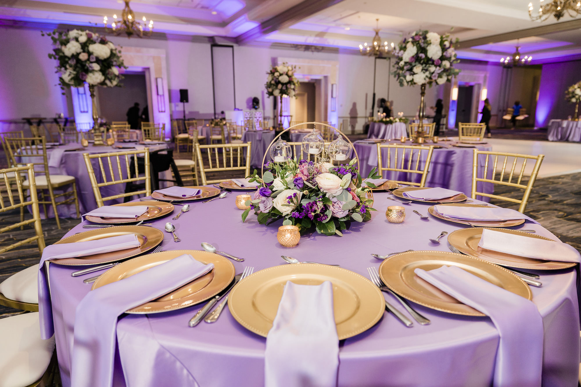 Gold Chargers with Lavender Satin Tablecloth and Napkin Linen | Silver Flatware | Round Arch with Violet, Pink, and White Flowers with Greenery Centerpiece Inspiration | Tampa Bay Rentals Gabro Event Services