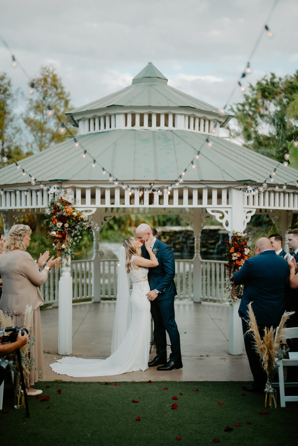 Bride and Groom First Kiss After Vows Under Outdoor Wedding Ceremony with White Gazebo and White Folding Chairs | Sarasota Florist Beneva Weddings