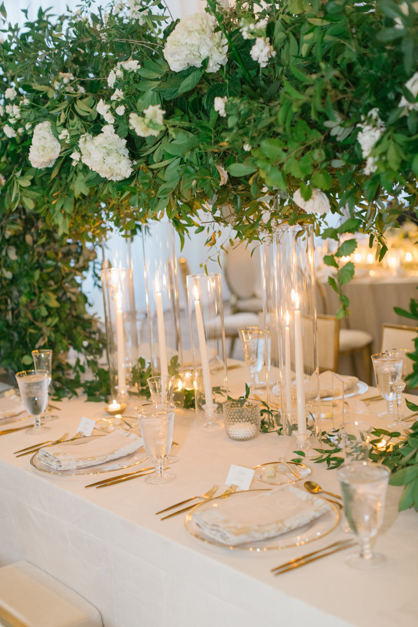 Luxurious Ballroom Wedding Reception Inspiration | Champagne Linen | Gold Rimmed Charger with Gold Flatware | Tabletop Arch White Flowers with Greenery Centerpiece Ideas | Hurricane Taper Candle Holders