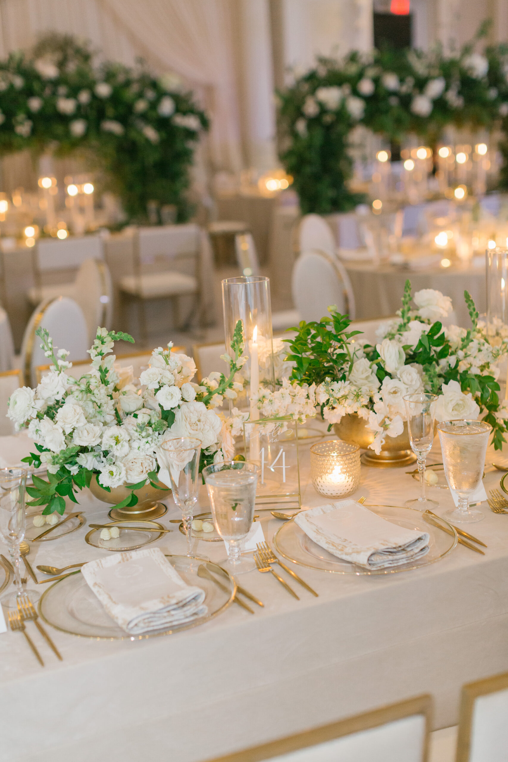 Luxurious Ballroom Wedding Reception Inspiration | Champagne Linen | Gold Rimmed Charger with Gold Flatware | White Flowers with Greenery Centerpiece Ideas | Hurricane Taper Candle Holders