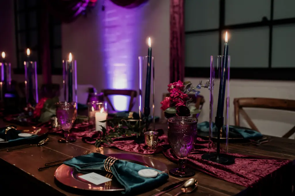 Dark and Moody Wedding Tablescape Inspiration with Black Pillar Candles, Gold Chargers, and Emerald Green Napkins and Purple Velvet Table Runner
