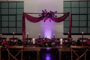 Dark and Moody Wedding Reception Sweetheart Table Inspiration with Deep Purple Velvet Draping and Black Pillar Candles on Wooden Feasting Farm Table