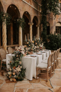 Feasting Table with Boho Wooden Rustic Style Chairs and Pastel Muted Floral Greenery Runner Centerpiece | Intimate Garden Floral Tablescape with Greenery and Bistro Lighting | Reception Decor Ideas